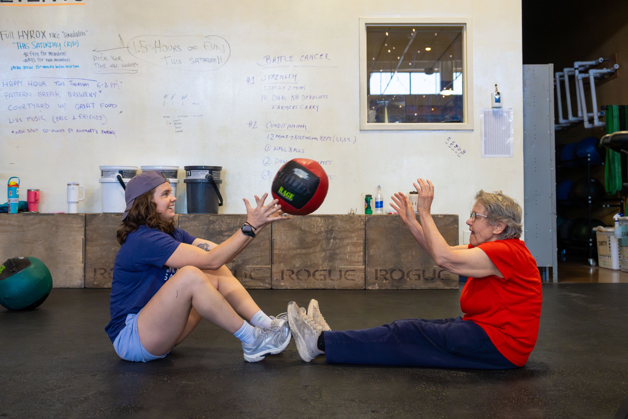 Two participants seated on the floor passing a medicine ball, both smiling